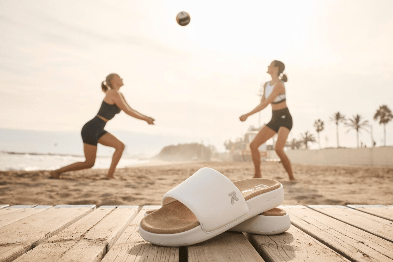 Rieker Damen Slides Narooma w3250-80 stehen am Strand, im Hintergrund spielen Frauen Volleyball.